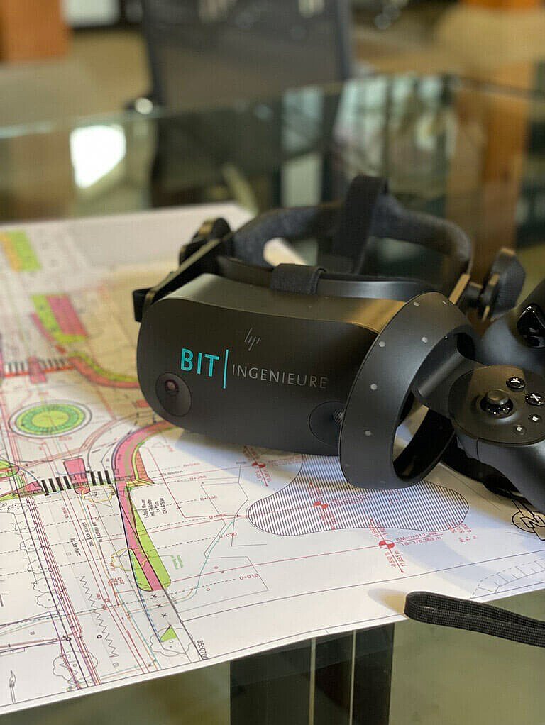 Eine VR-Brille mit dem Logo BIT Ingenieure liegt auf einem Tisch und Bauplänen.